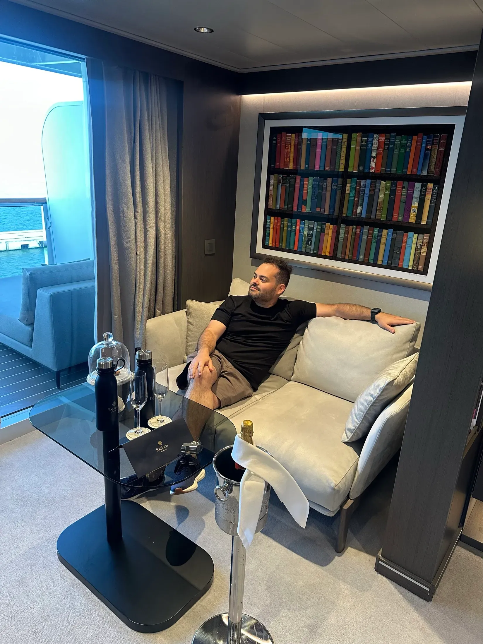 Me on the sofa of my Explora I suite, the library-spine artwork and the ocean terrace behind me